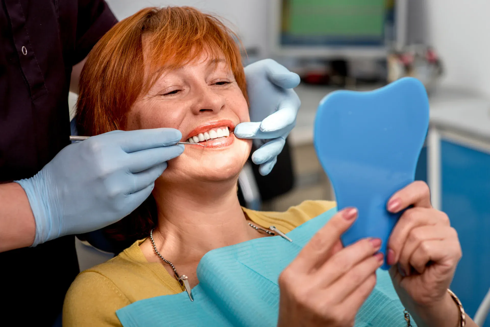 Patient smiling while dentist checks her teeth as she holds a tooth-shaped mirror